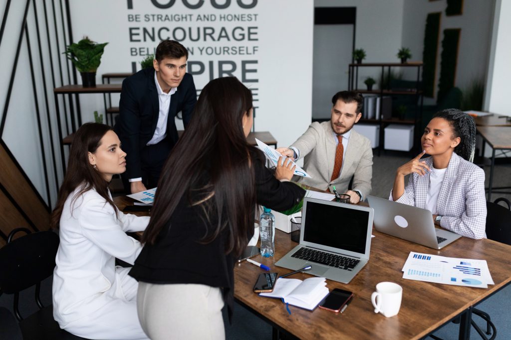 Grupo de vendedores calculan comision de ventas están sentados en una mesa cuadrada de madera con papeles y gráficos sobre ella. Se pasan los unos a los otros papeles y tienen ordenadores sobre la mesa.